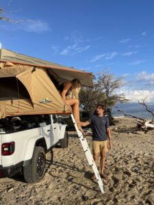 Happy couple enjoying a beach day next to a Jeep camper with a rooftop tent on the sandy shores of a beach on Maui.